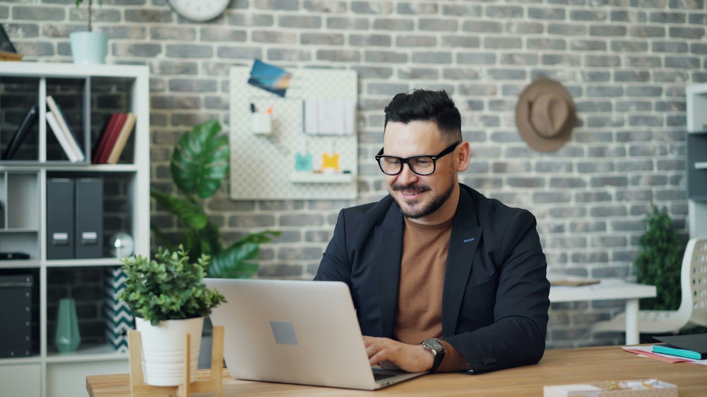 Un homme barbu sourit en travaillant sur un ordinateur portable à un bureau.